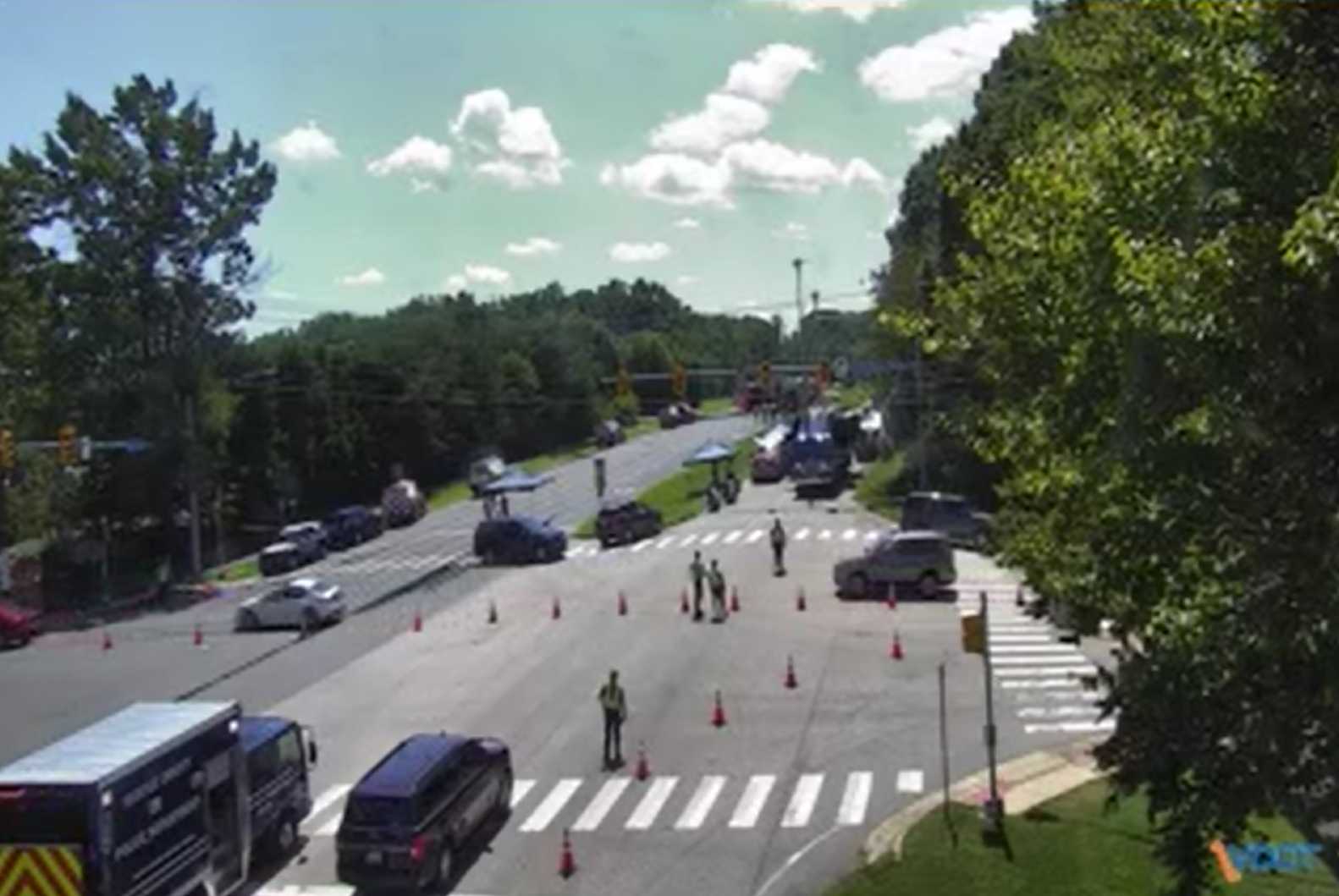 Busy suburban street closed for roadwork with orange cones, workers guiding traffic, and parked cars under a blue sky with trees on both sides.