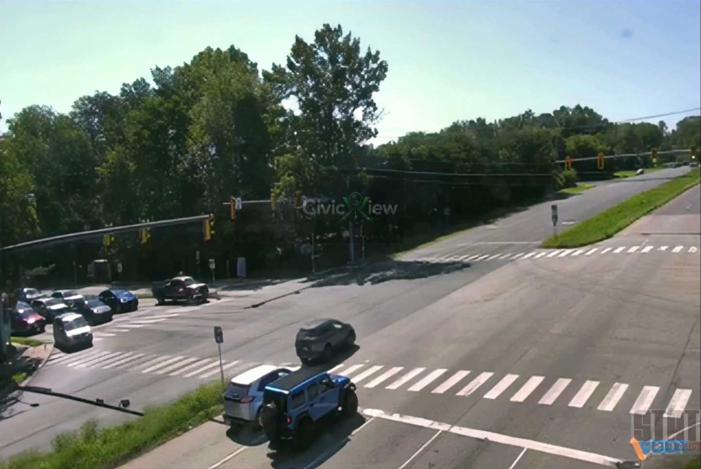 Wide view of a multi-lane intersection with zebra crosswalks, traffic lights, and several parked and moving cars; trees line the far side of the street at noon sun.