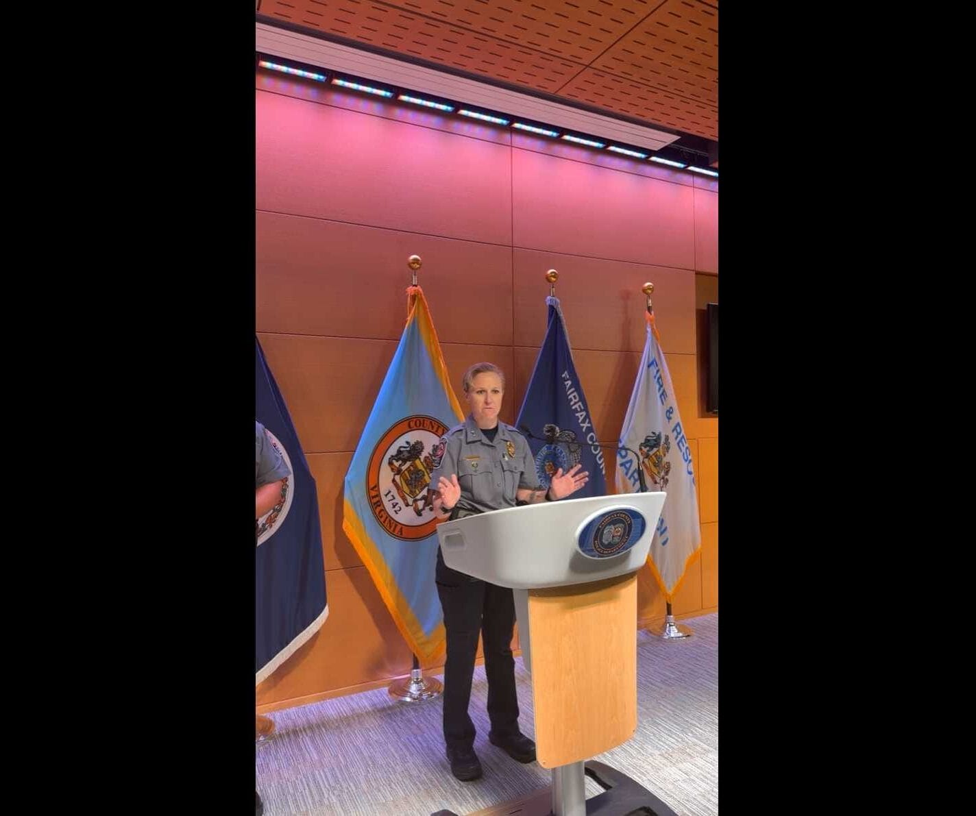 Female officer in uniform standing at a lectern with multiple flags behind her, delivering a speech in a conference room