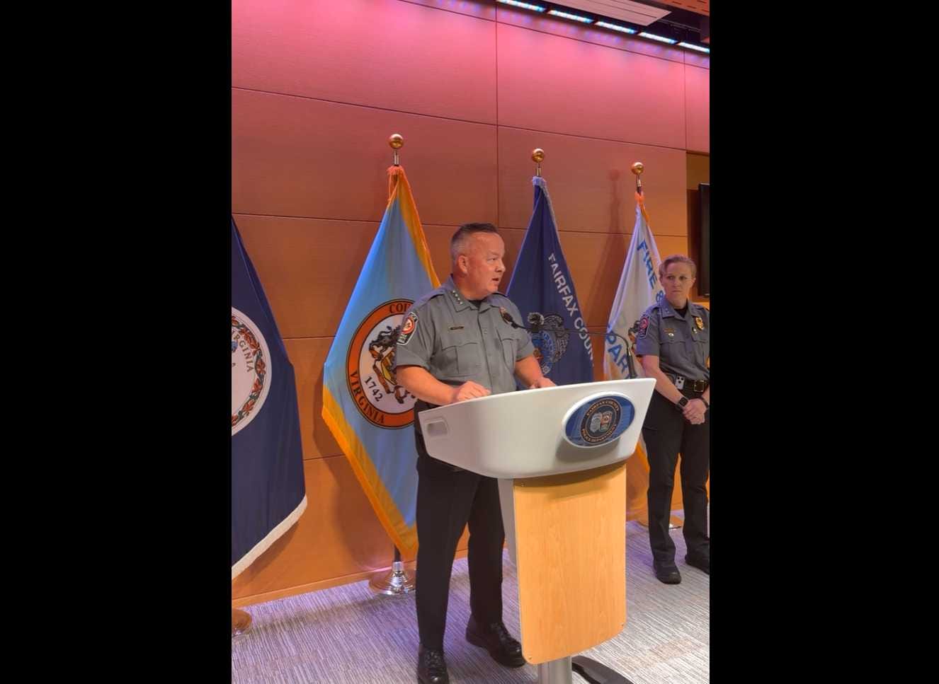 Fairfax County Police Chief Kevin Davis at an Aug. 19, 2026 press conference about the fatal stabbing of Carmen Puch in Great Falls (via FCPD/Facebook) | FFXnow Uniformed man speaking at a podium with a seal, as flags stand behind him; another officer stands nearby in a briefing or ceremony setting.