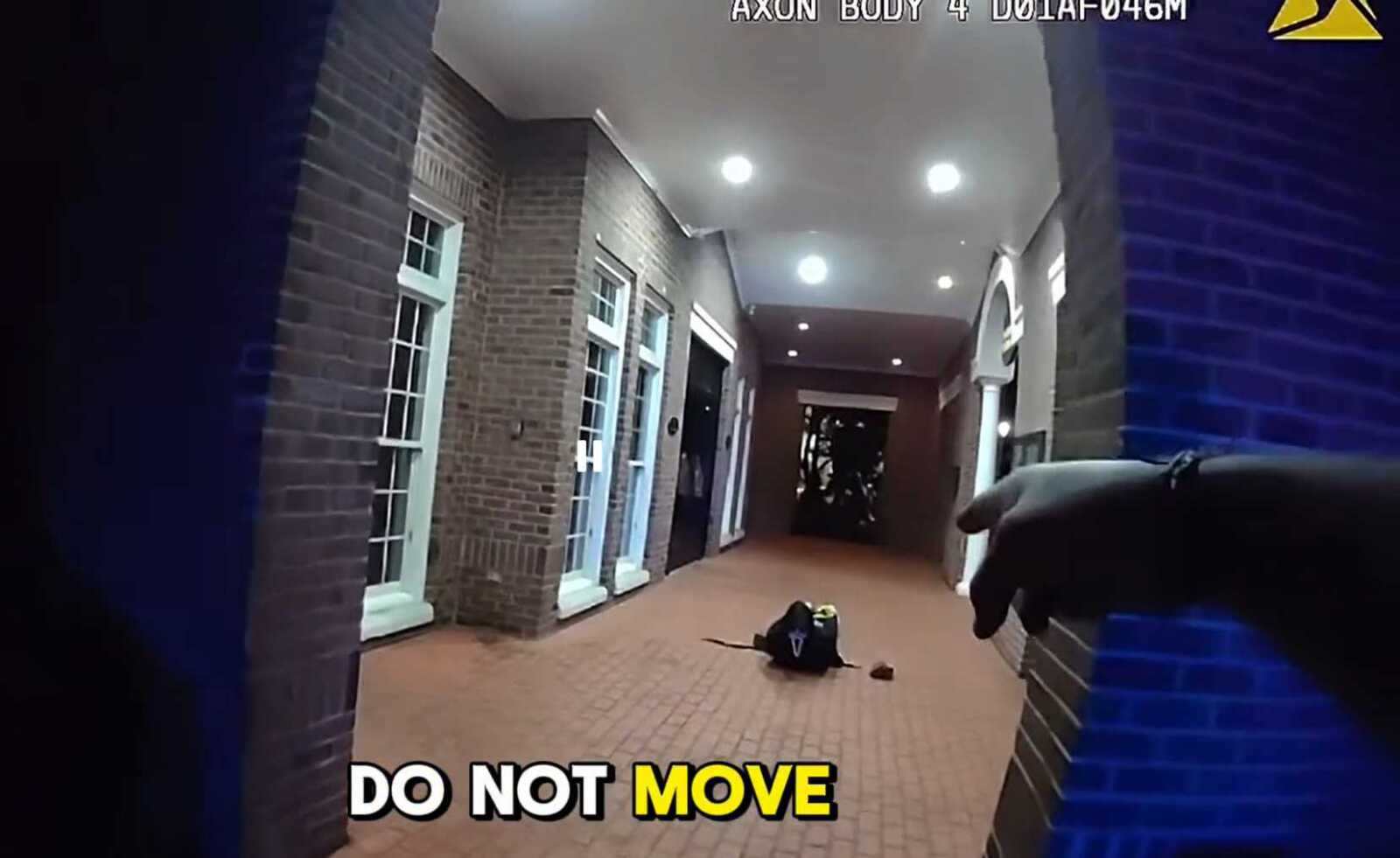 First-person view from a body-worn camera in a brick hallway; a person lies on the floor ahead while a gloved hand points from the right; caption reads 'DO NOT MOVE'.