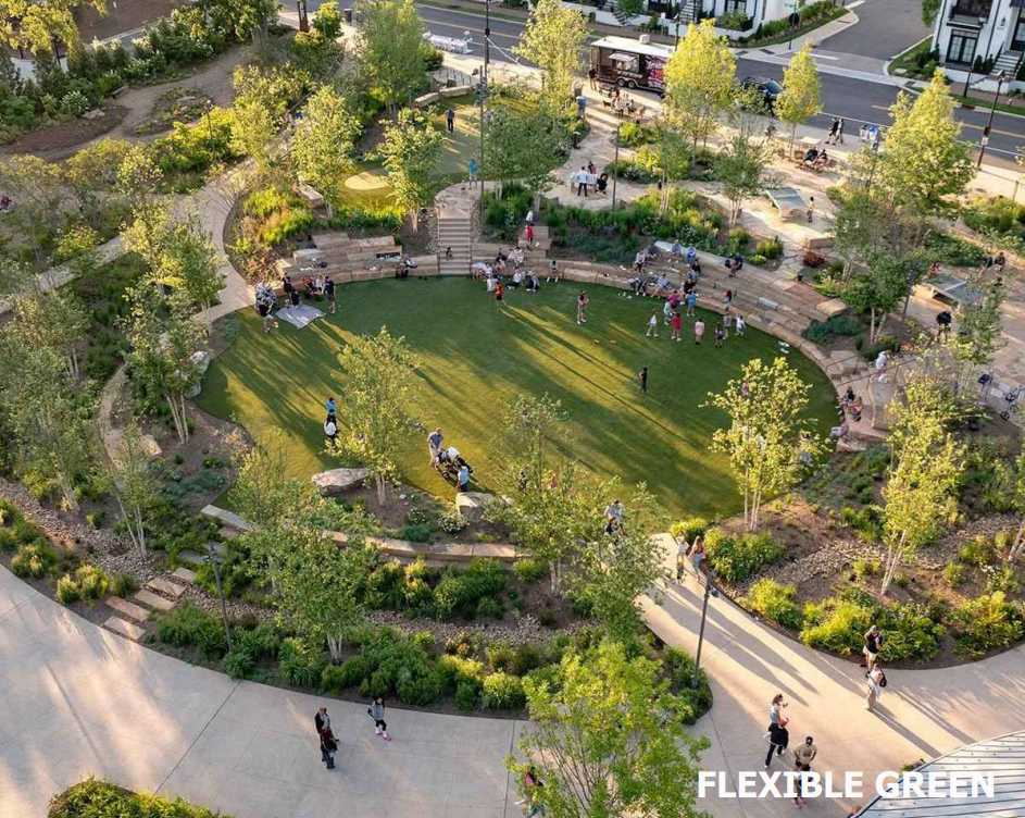 Aerial view of a circular green park with terraced seating, surrounding trees, and people walking or sitting around the lawn in an urban area.