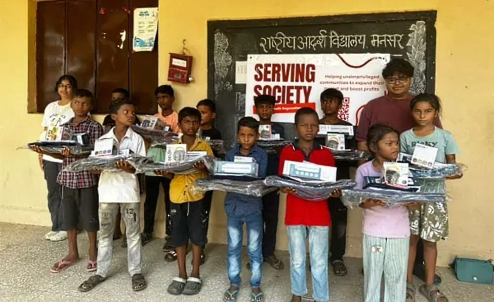Serving Society Global Foundation supports initiatives in India (courtesy SSGF) | FFXnow Group of children standing in a line, each holding wrapped packages and books donated for a charity event in front of a 'SERVING SOCIETY' banner.