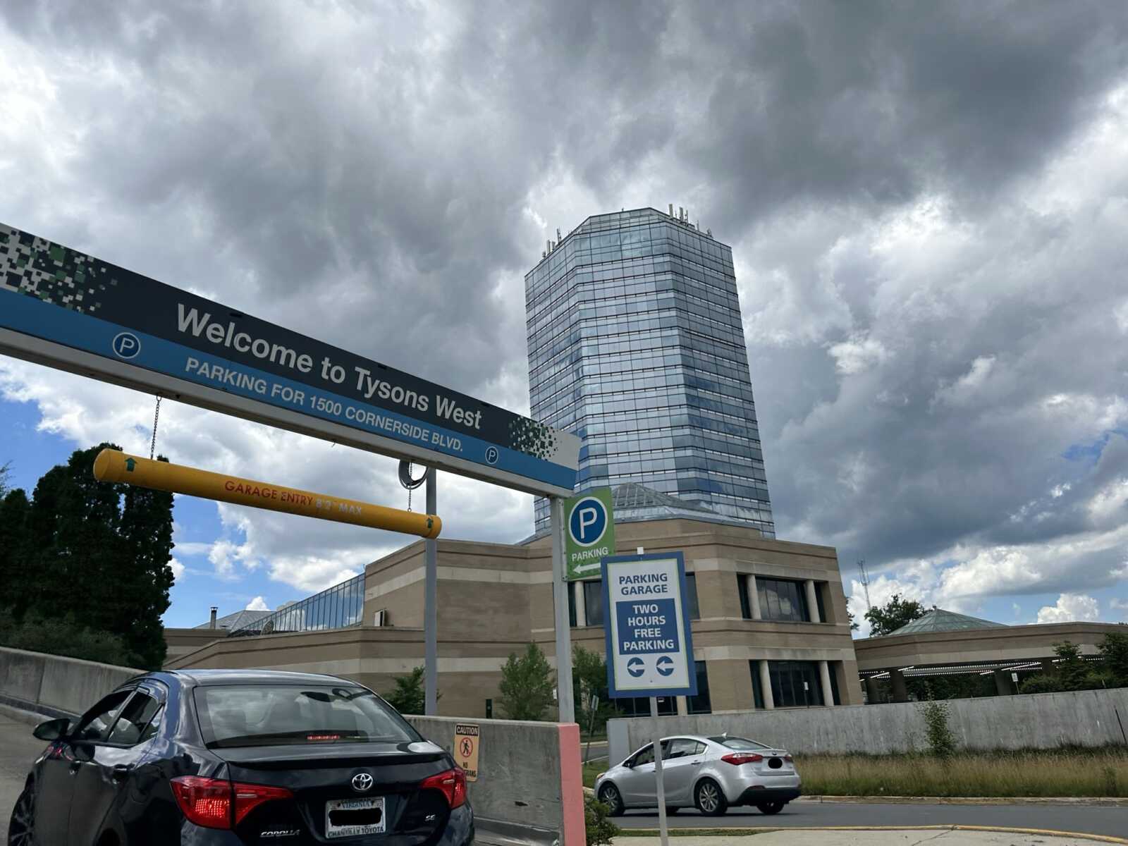 Welcome to Tysons West sign at a parking entrance with a glass high-rise building in the cloudy sky behind it.