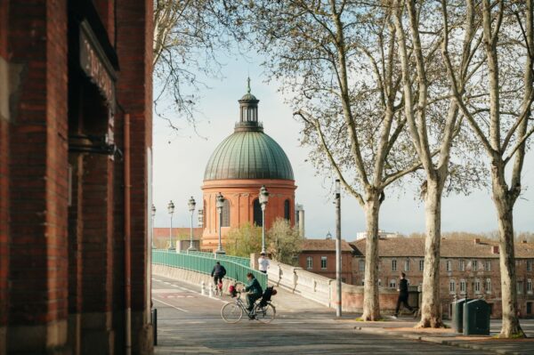 Red brick street scene with a green-domed historic building in the background, framed by trees and a pedestrian walkway with cyclists and walkers in the foreground.