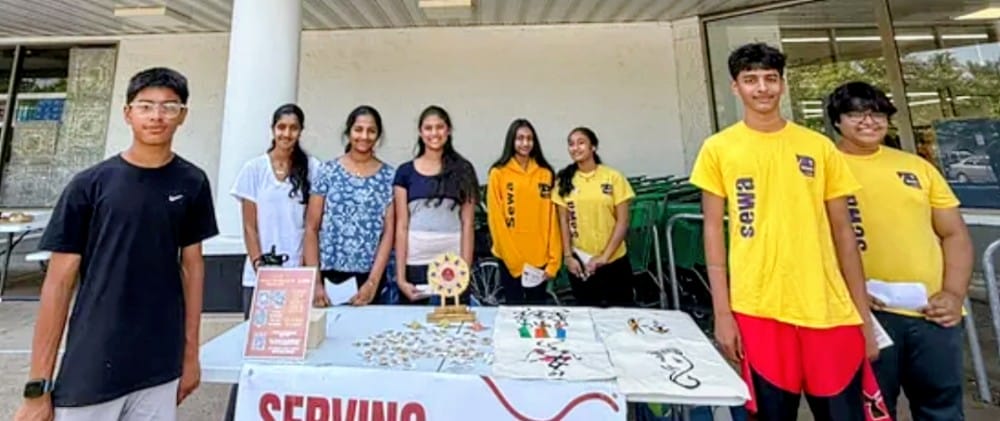 Volunteers from the the SEWA DC Youth Group support the Serving Society Global Foundation (courtesy SSGF) | FFXnow Group of students posing behind a table at a campus booth with display items and signs outside a building.