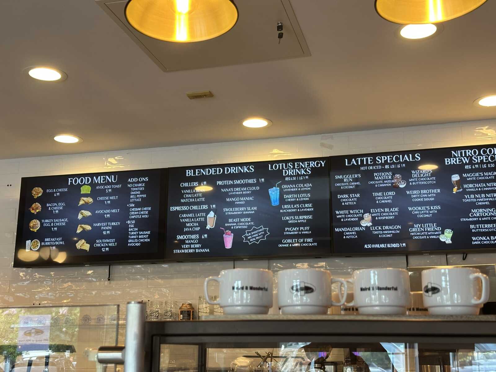 Cafe menu boards with food and drink listings under warm ceiling lights; cups visible in foreground.