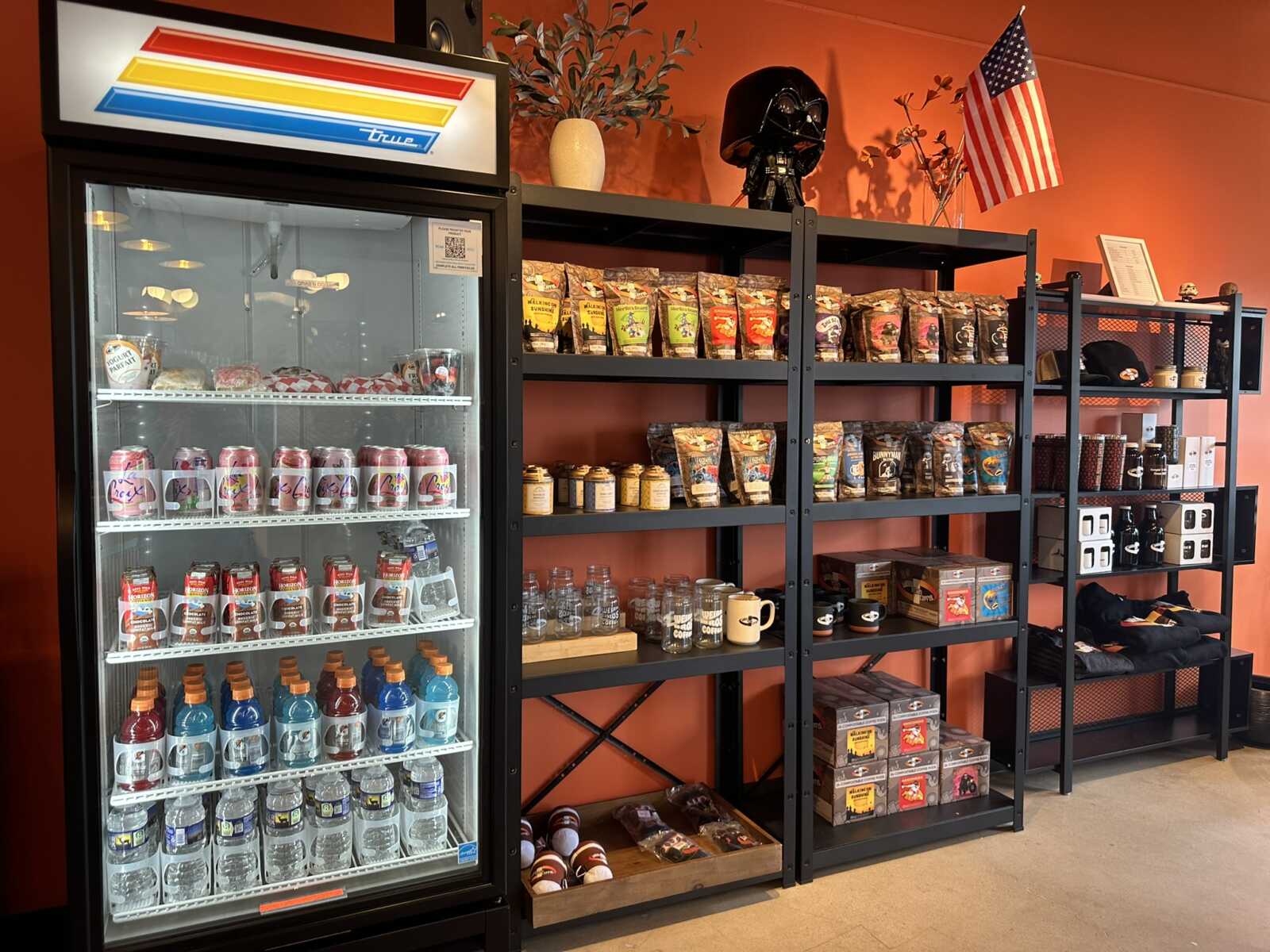 Glass‑front cooler on the left overflows with bottles; black metal shelving on the right holds bags of snacks, mugs, and merchandise atop an orange wall with an American flag and a Darth Vader helmet.