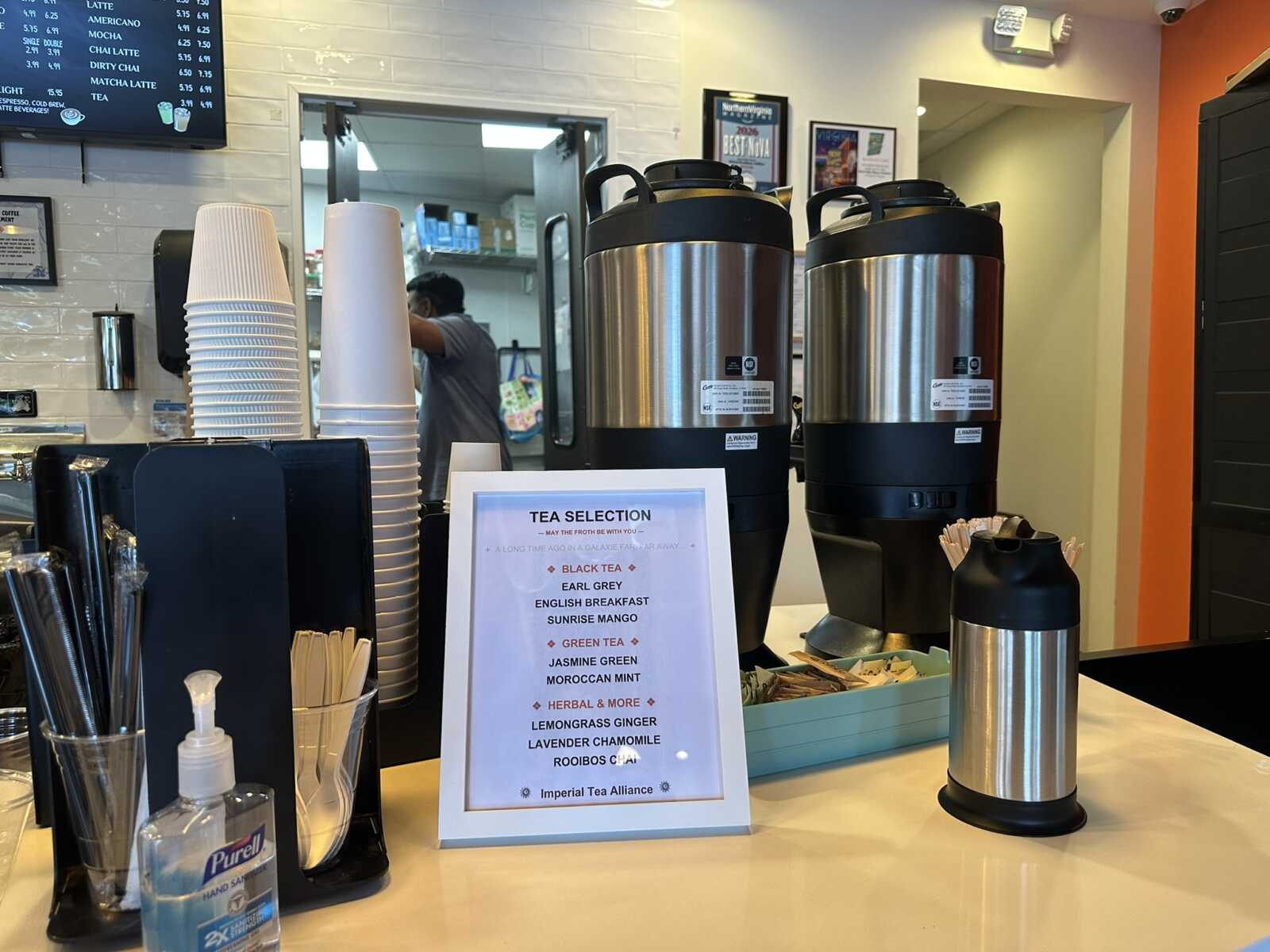 Cafe counter with two large stainless steel urns, plastic cups, and a framed Tea Selection menu on the counter.