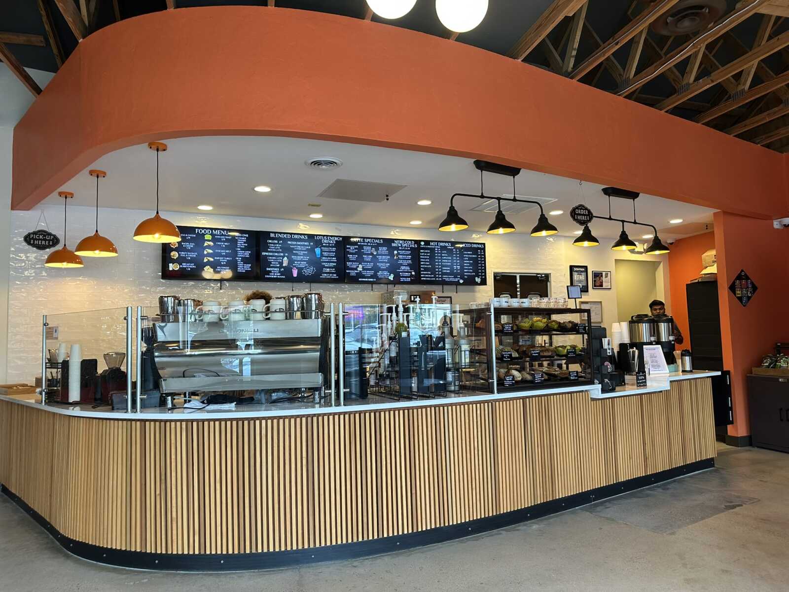 Cafe interior with curved orange wall above a long wooden counter and glass display case of pastries and equipment behind the bar.