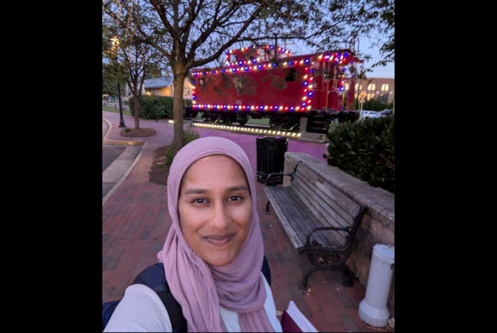 Yasmin Shafiq in front of Herndon's caboose (courtesy Yasmin for Herndon) | FFXnow Selfie of a woman in a pink hijab smiling in a park with a red decorated train/float in the background.