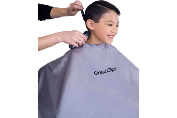 Boy smiling while a barber trims his hair with clippers, wearing a gray Great Clips cape.