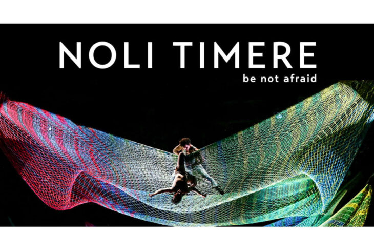 Banner for 'NOLI TIMERE' (be not afraid) with two performers on a colorful net sculpture against a dark background, top-centered text.