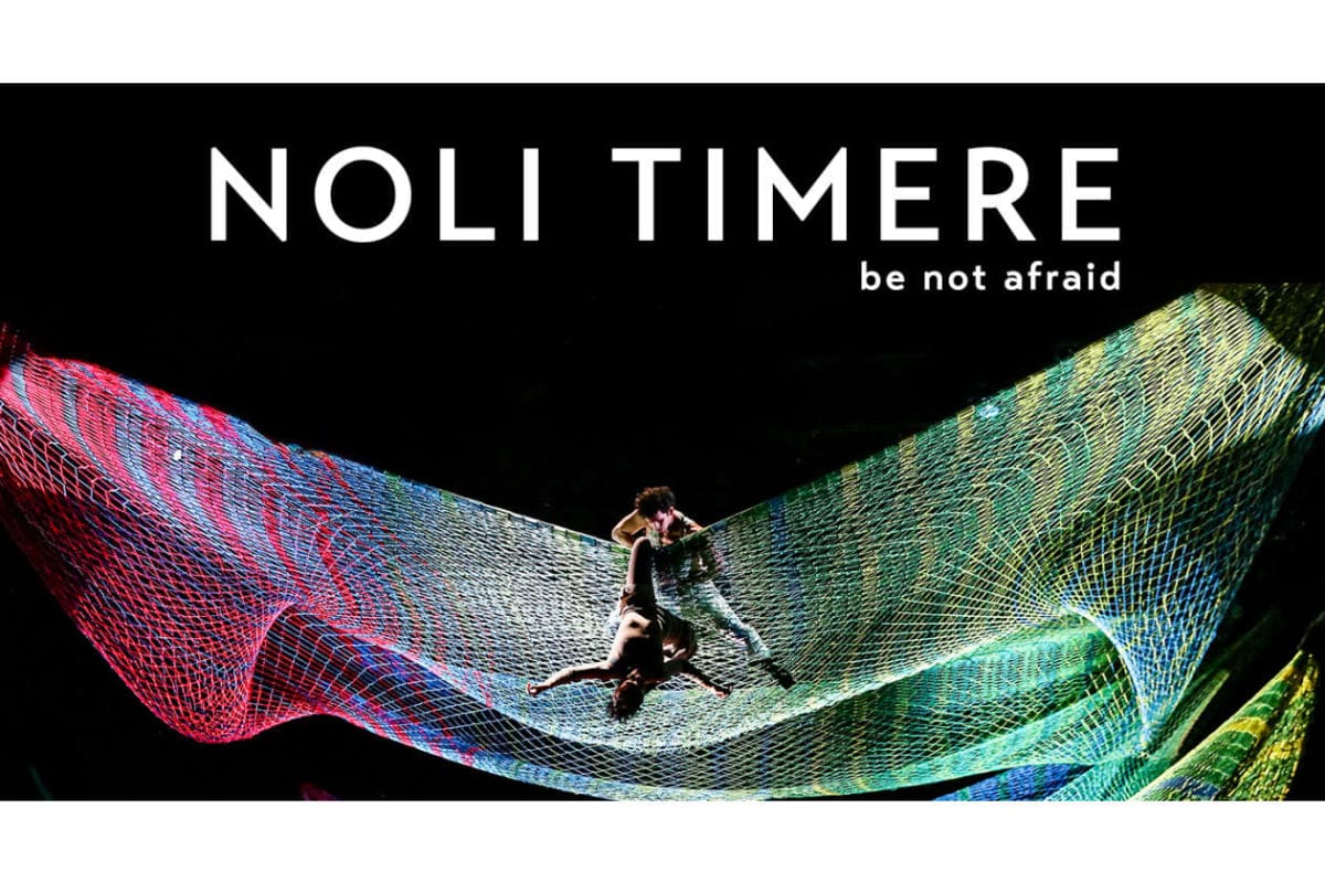 Banner for 'NOLI TIMERE' (be not afraid) with two performers on a colorful net sculpture against a dark background, top-centered text.