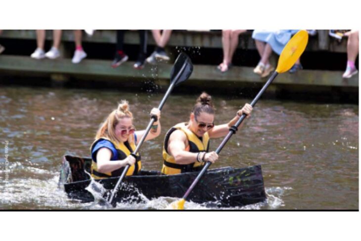 Two people in yellow life jackets paddle a black canoe on a river while spectators sit on a dock nearby.