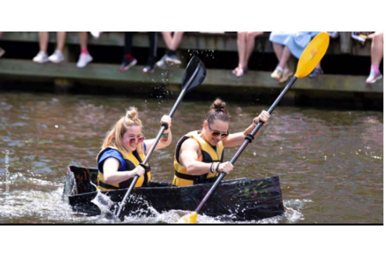Two people in yellow life jackets paddle a black canoe on a river while spectators sit on a dock nearby.