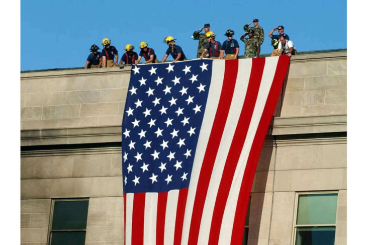 Group of people in helmets on a building rooftop lowering a large American flag down the side of the wall; clear blue sky behind them.