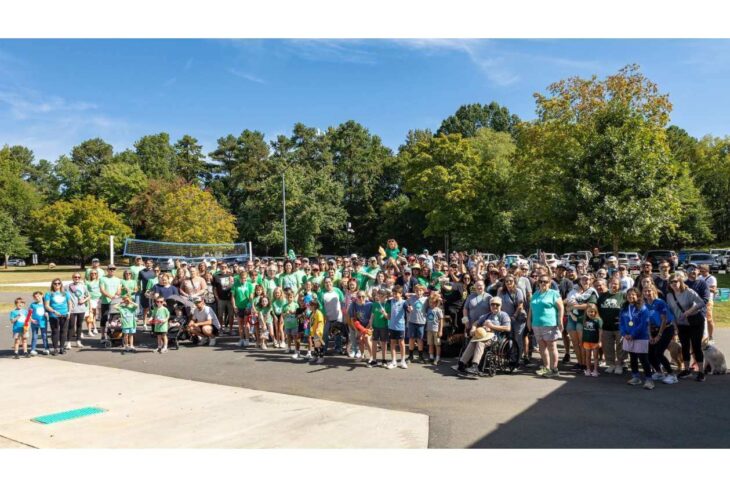 Large group of people gathered outdoors for a community event, many wearing green shirts in front of trees and a volleyball net.