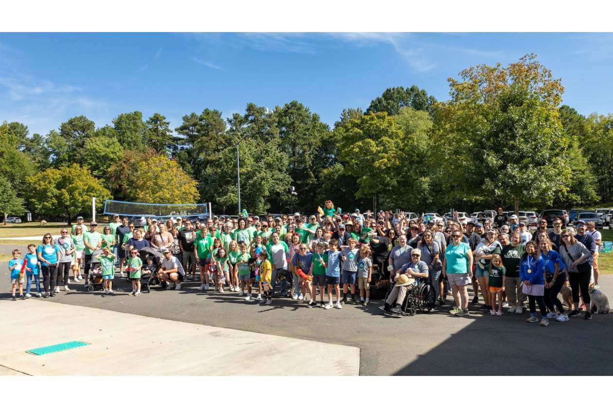 Large group of people gathered outdoors for a community event, many wearing green shirts in front of trees and a volleyball net.