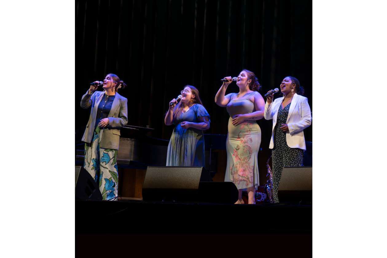 Four female vocalists sing into wireless mics on a dimly lit stage, wearing colorful outfits and standing in a row with a piano in the background.