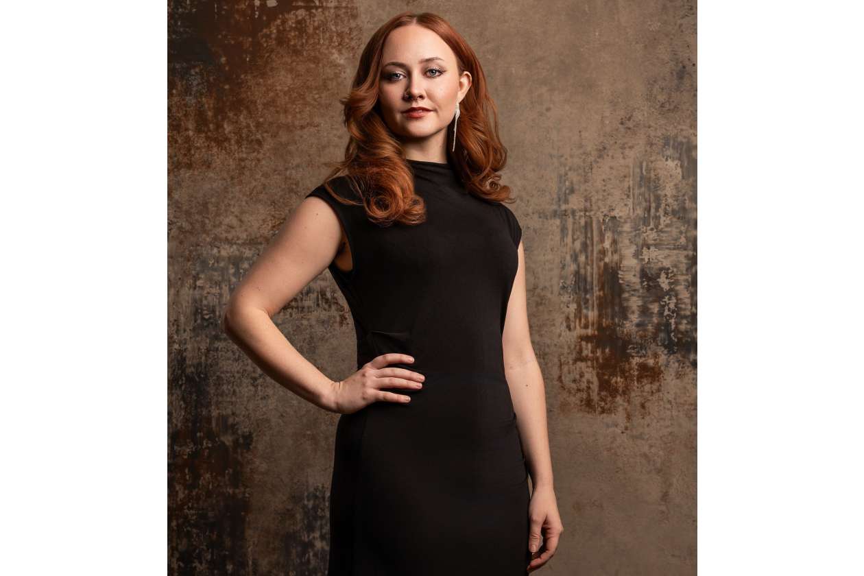 Woman in a black sleeveless dress posing with hand on hip against a rustic textured backdrop.