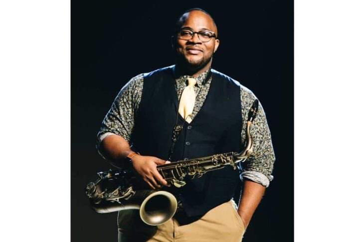 Smiling man in a black vest and patterned shirt holds a saxophone at his side, wearing glasses and a yellow tie.