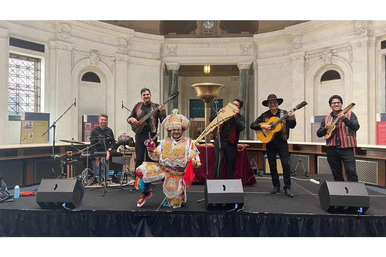 Live music performance in a grand marble hall; a dancer in bright, ornate costume kneels center while the band plays behind.