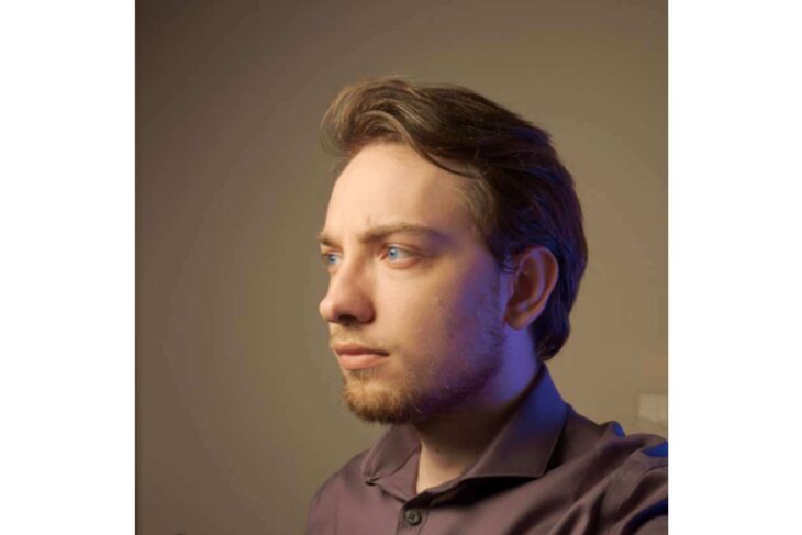 Side-profile portrait of a man with short brown hair and light stubble, wearing a purple shirt, lit by warm studio lighting.