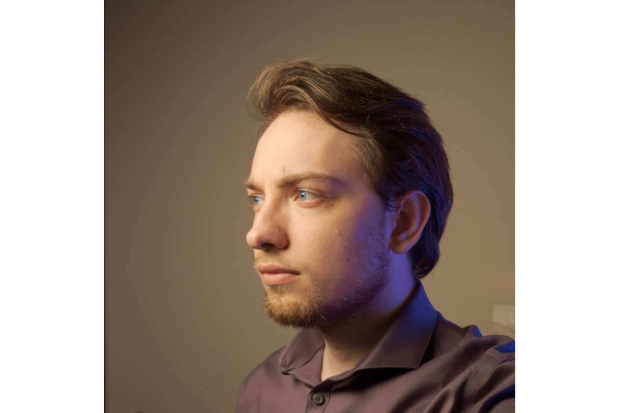Side-profile portrait of a man with short brown hair and light stubble, wearing a purple shirt, lit by warm studio lighting.