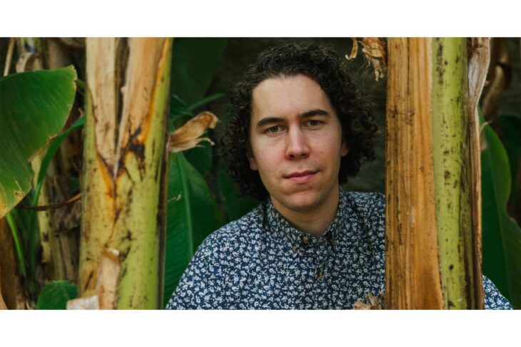 Portrait of a young man with curly hair standing among banana plants in a lush outdoor setting, facing the camera.