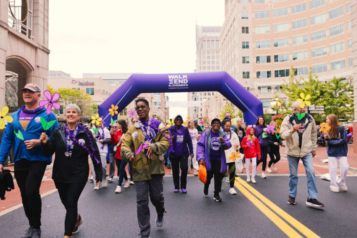 Crowd of diverse participants walking under a purple 'Walk to End Alzheimer's' arch on a city street, holding colorful flower props and wearing purple attire.