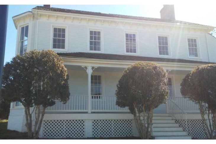 Two-story white house with a front porch, lattice skirting, and trimmed shrubs in the yard.