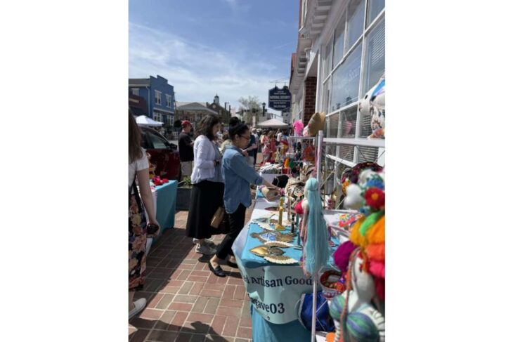 Outdoor street market with shoppers browsing a table of handmade crafts and colorful decorations outside storefronts on a sunny day