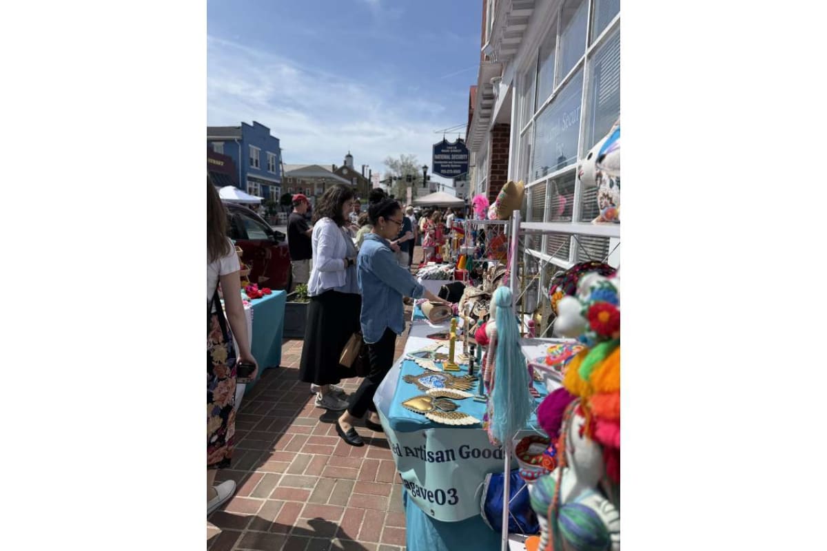 Outdoor street market with shoppers browsing a table of handmade crafts and colorful decorations outside storefronts on a sunny day