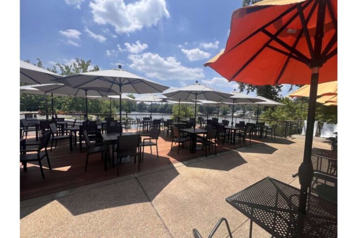 Outdoor patio with numerous black tables and chairs under large white umbrellas by a lake on a sunny day