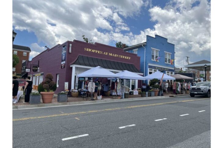 Outdoors market on a sunny day with white tents along a brick storefront row on Main Street; shoppers browse clothing and goods.