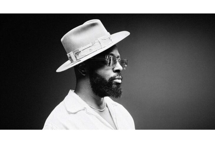 Black-and-white portrait of a man with a beard wearing a wide-brim hat, sunglasses, an open shirt, and a pearl necklace against a dark gradient background.