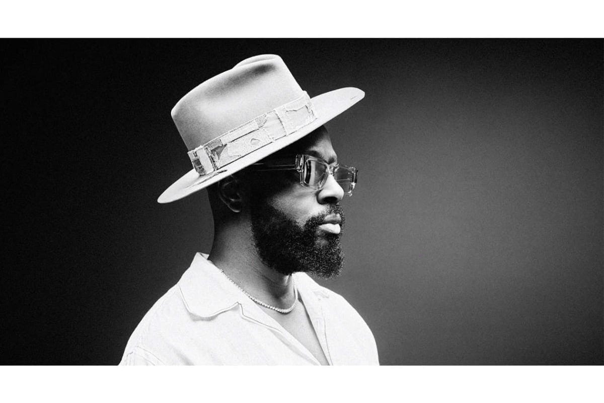 Black-and-white portrait of a man with a beard wearing a wide-brim hat, sunglasses, an open shirt, and a pearl necklace against a dark gradient background.