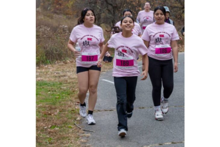 Group of women in pink T-shirts running together in a 5K, bib numbers 186–189 visible on a paved park path.
