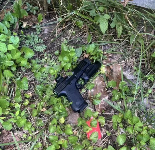 A black pistol lies on dirt among green groundcover and grass, with a red rectangular item nearby.