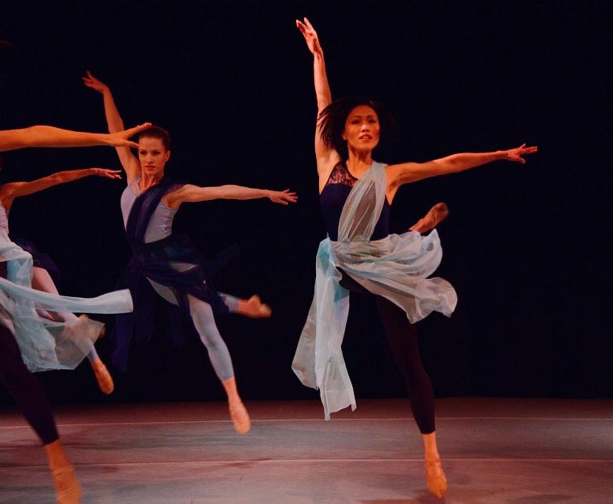 Dancers in blue costumes perform a ballet routine on stage, turquoise scarves swirling.