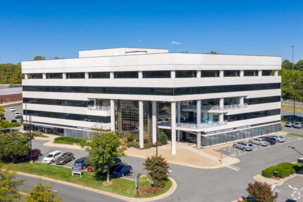 A modern white office building with glass windows, three stories tall, surrounded by a parking lot and greenery (aerial view).