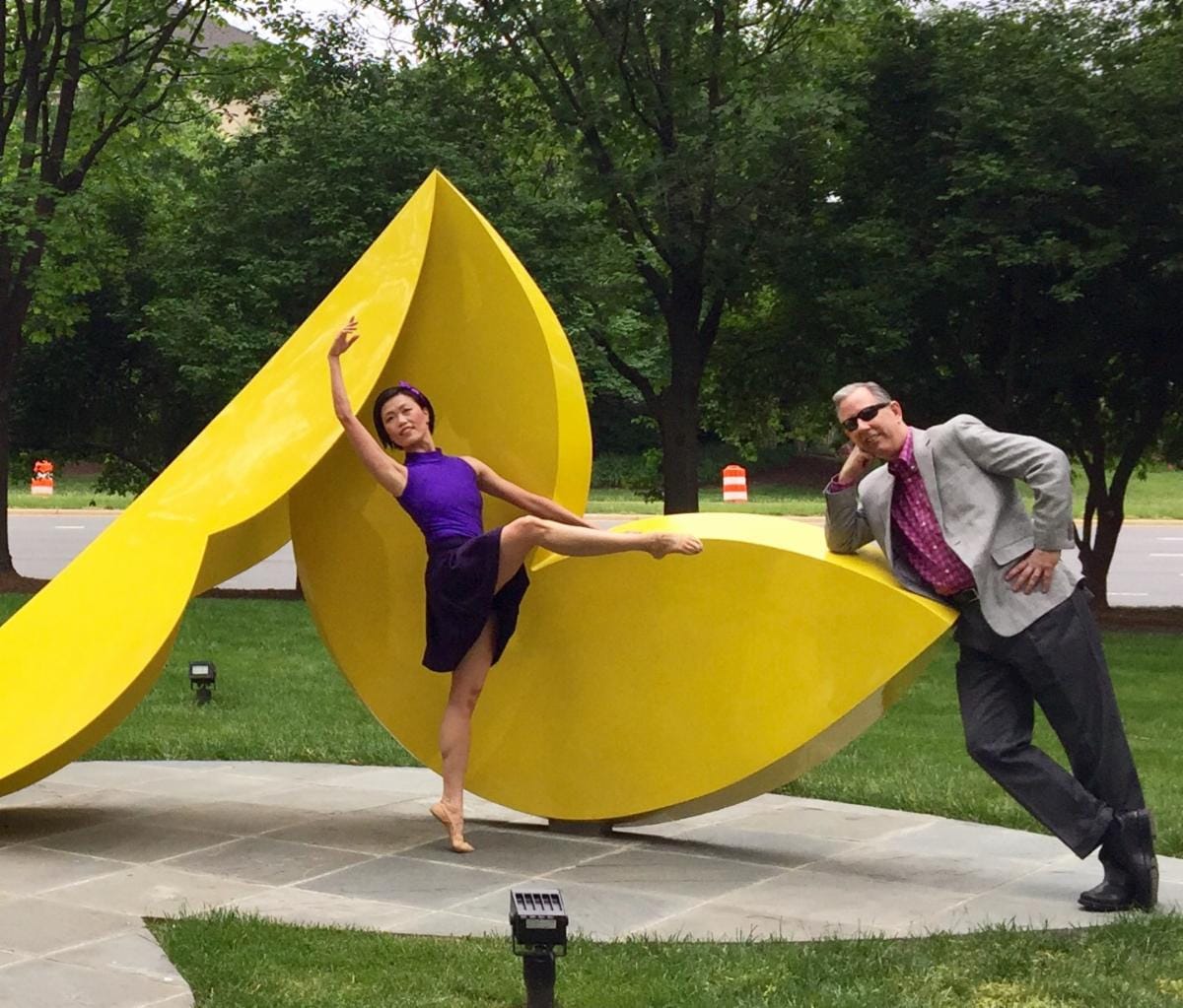 A ballerina in a purple leotard dances on a bright yellow abstract sculpture outdoors, while a man in a gray blazer leans on the sculpture and smiles nearby.