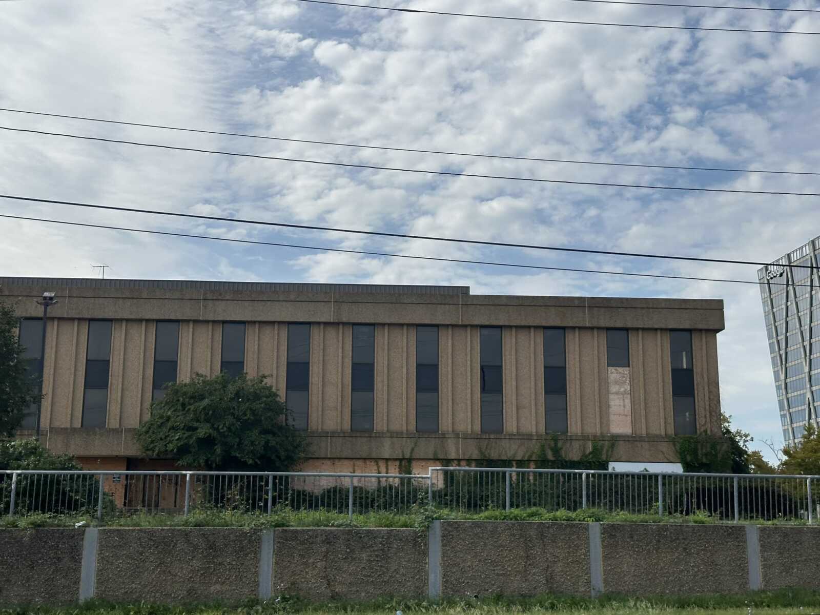 Beige brick office building with tall vertical windows, a front fence and shrubs, set against a cloudy sky with power lines crossing overhead.