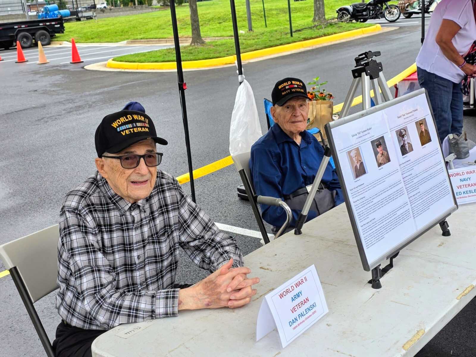 Two elderly World War II veterans wearing hats sit at a table with display boards about their service in a parking lot outside.