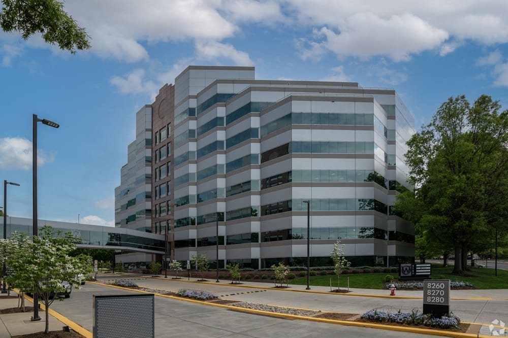 Modern multi-story office building with glass windows and a blue sky backdrop, surrounded by a parking area and landscaped planters.