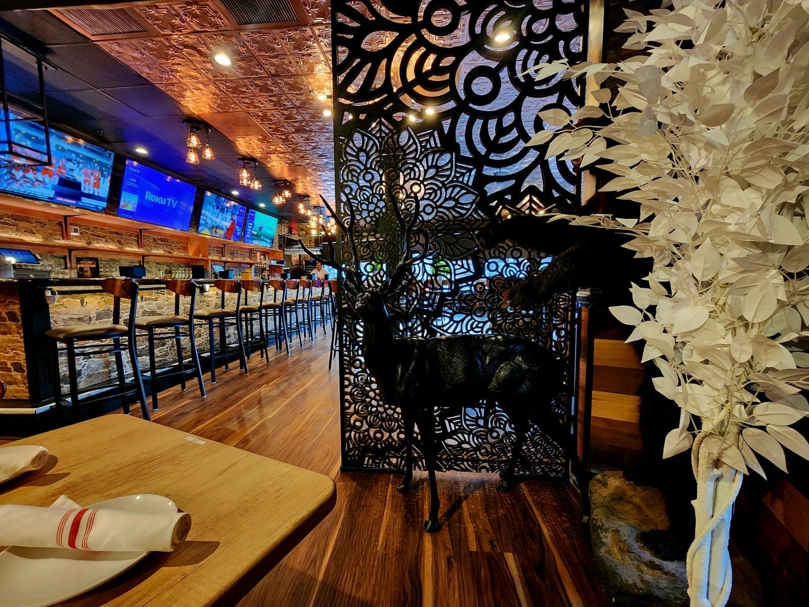 Restaurant interior with a long bar, stools, and several TVs above the counter; a black decorative screen with a deer silhouette stands beside a white leafy plant.