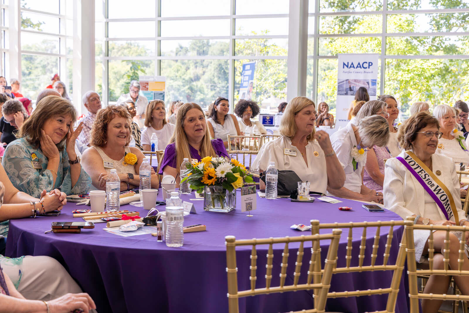 Group of women seated at a round table with a purple tablecloth, flowers, and water bottles at a formal event, listening attentively.