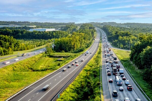 Aerial view of a multi-lane highway surrounded by green trees, with heavy traffic on the right and lighter traffic on the left lane Waves into the distance under a blue sky.