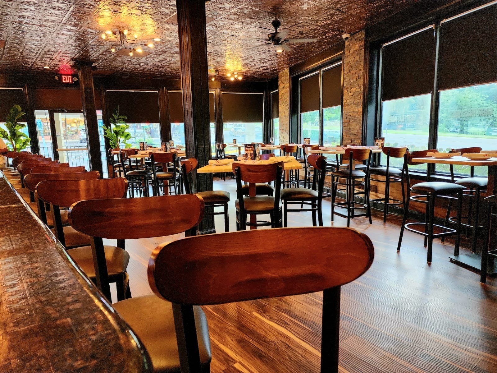 Empty rustic restaurant interior with dark wooden chairs and high tables along large windows, warm pendant lighting, and exposed tin ceiling.